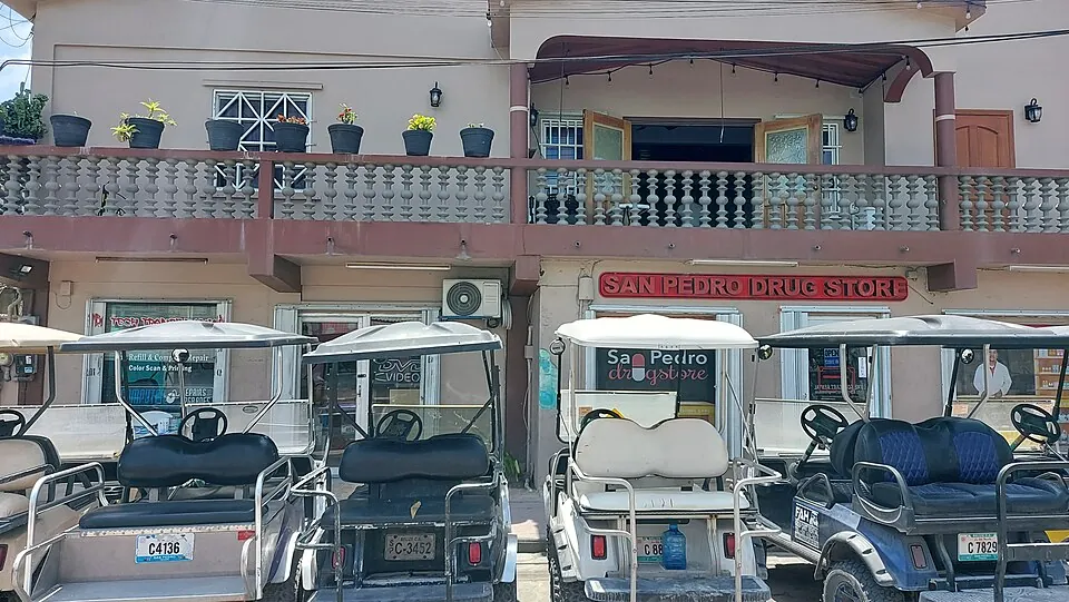 Four golf carts with Belize licence plates parked in a row in front of the San Pedro Drug Store, a two-storey building on a San Pedro street.