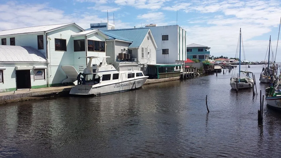 A water taxi lettered Caye Caulker Water Taxi moored alongside waterfront buildings in Belize City, with sailing boats on the channel behind.