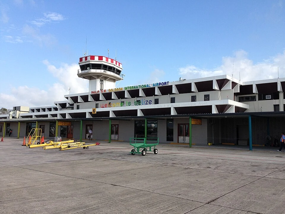The terminal building at Philip S. W. Goldson International Airport, its control tower behind and the airport name and the words welcome to belize lettered across the facade, seen from the apron.