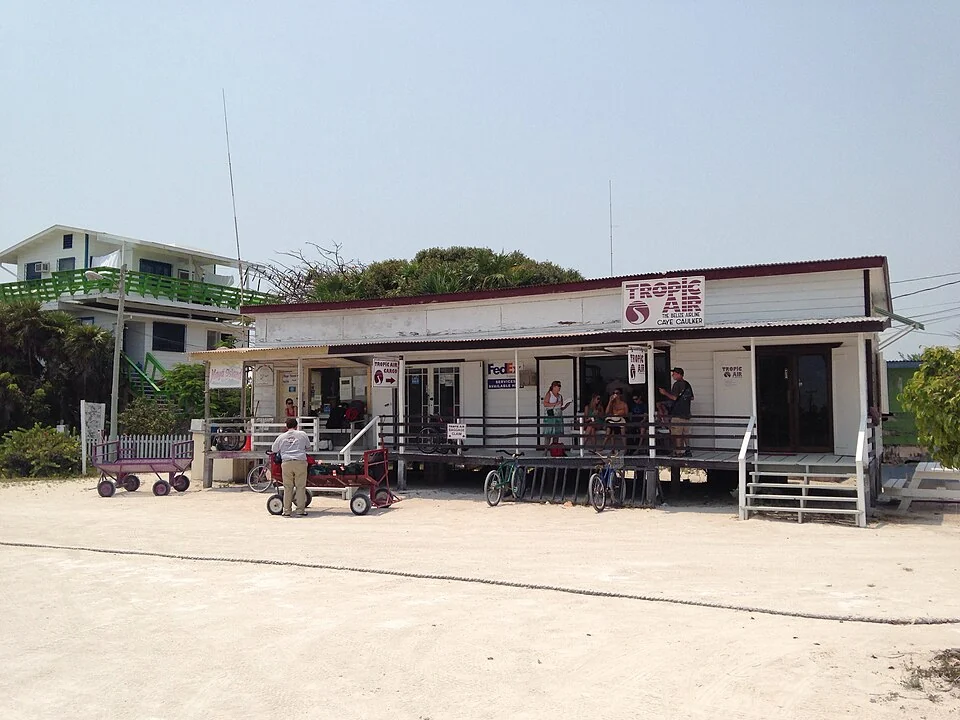 A low white timber building at the Caye Caulker airstrip with a Tropic Air sign on its roof, passengers waiting on the veranda and handcarts and bicycles parked on the sand outside.