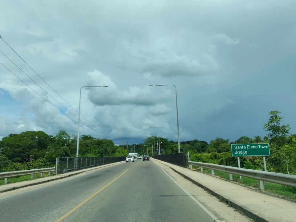 The highway crossing the Santa Elena Town Bridge towards San Ignacio, two lanes of traffic ahead and a green road sign reading Santa Elena Town Bridge on the right.