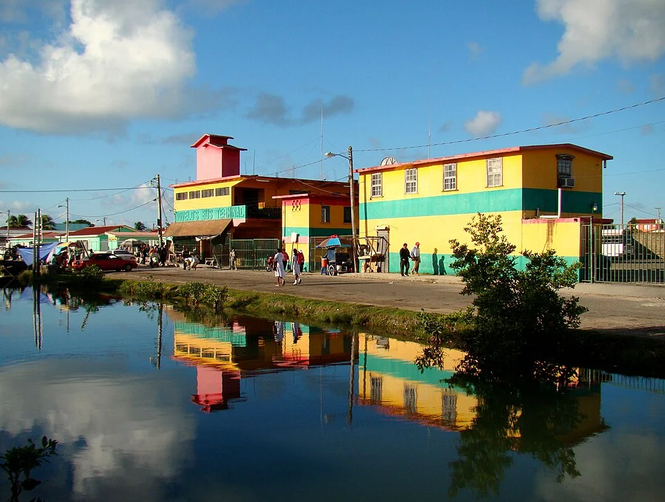 A two-storey building painted in yellow, green, red and pink lettered Novelo's Bus Terminal, seen across a channel of still water in Belize City with people walking outside it.