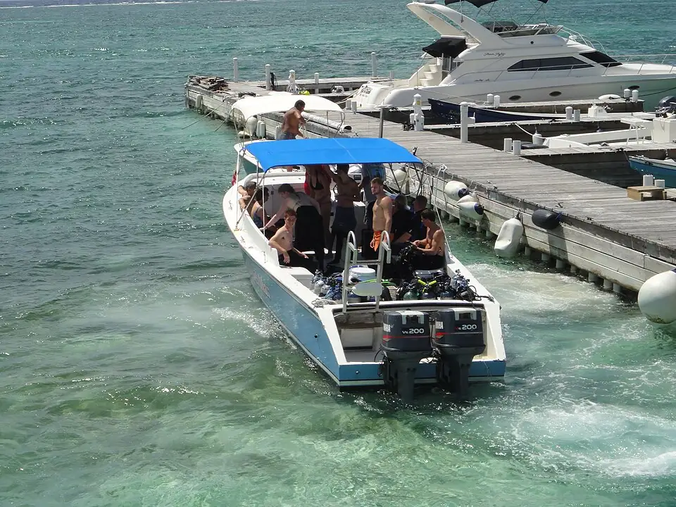 A dive boat carrying divers and scuba tanks pulling away from a wooden dock in clear shallow water, with a larger boat moored alongside.