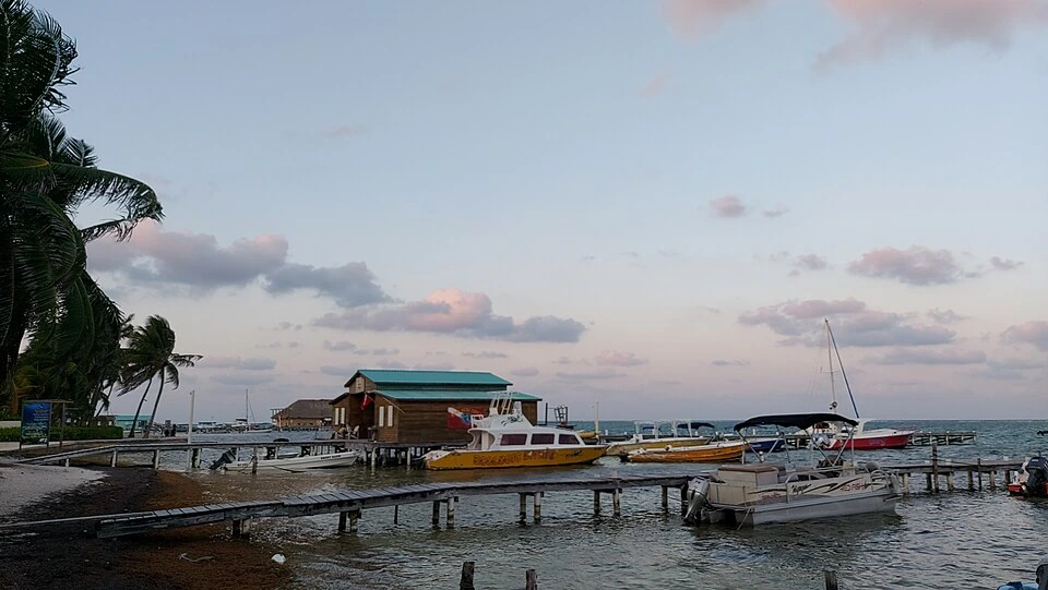 Dive boats moored along wooden jetties in front of a stilted timber dive centre with a green roof, palms on the shore and a pale evening sky behind.
