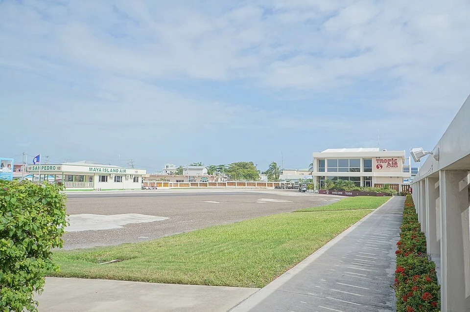 An empty airstrip apron under a blue sky, with a low green Maya Island Air terminal on the far side and a white Tropic Air terminal building on the near side.
