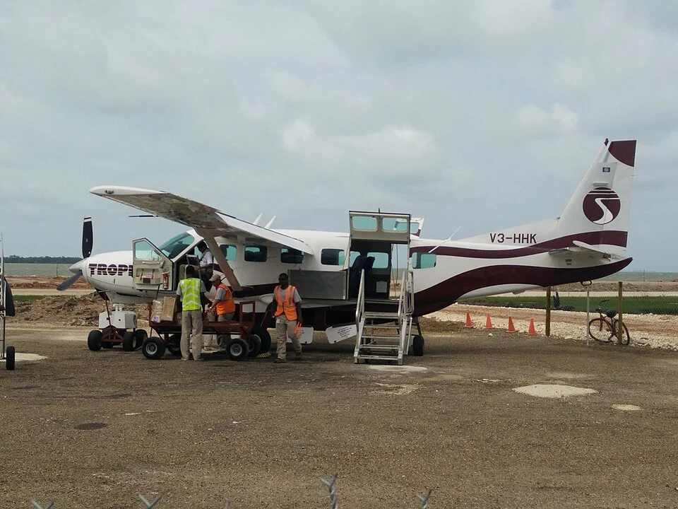 Ground staff in high-visibility vests loading boxes and bags from a trolley into the hold of a single-engine Cessna Caravan parked on a gravel apron.