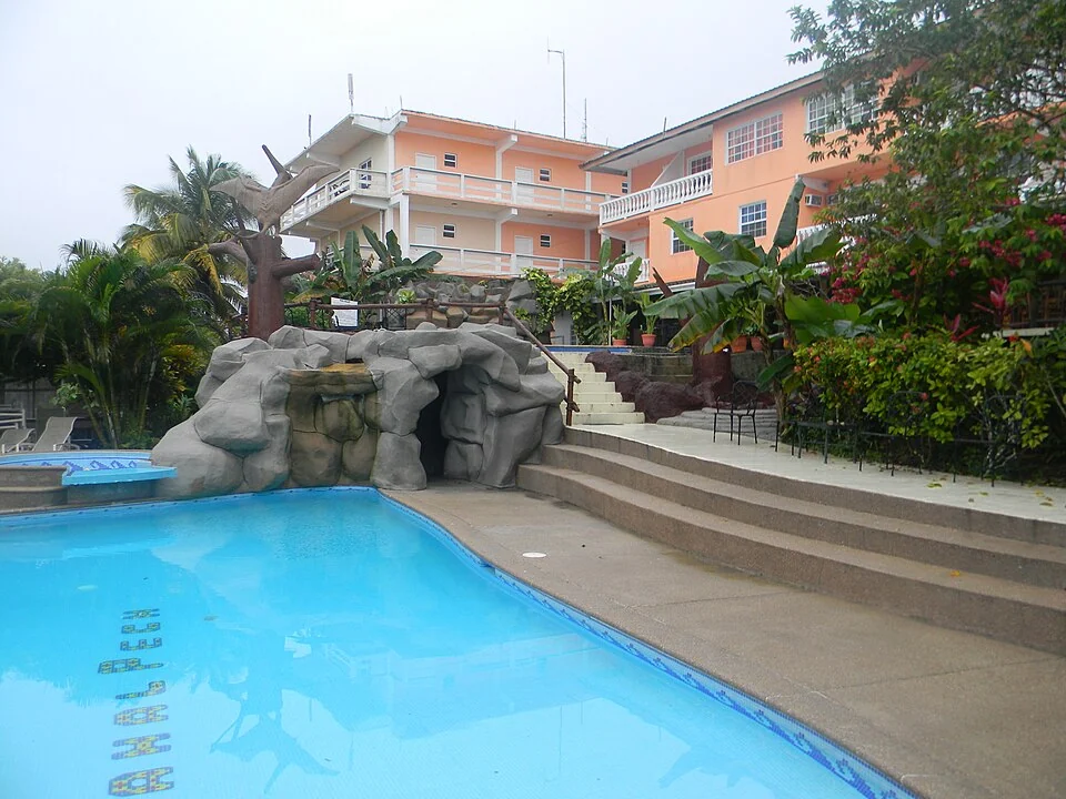 A resort swimming pool with a moulded rock feature at one end, backed by two peach-painted guest buildings with white balconies and tropical planting.