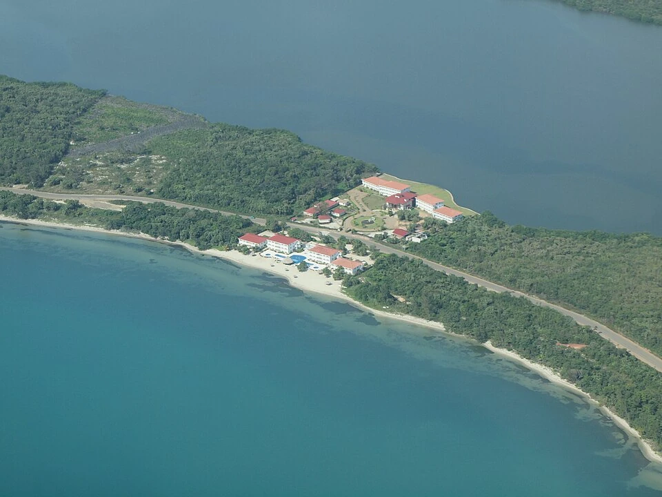 An aerial view of a narrow forested peninsula between two stretches of sea, with a cluster of red-roofed resort buildings and a pale beach along the seaward side.