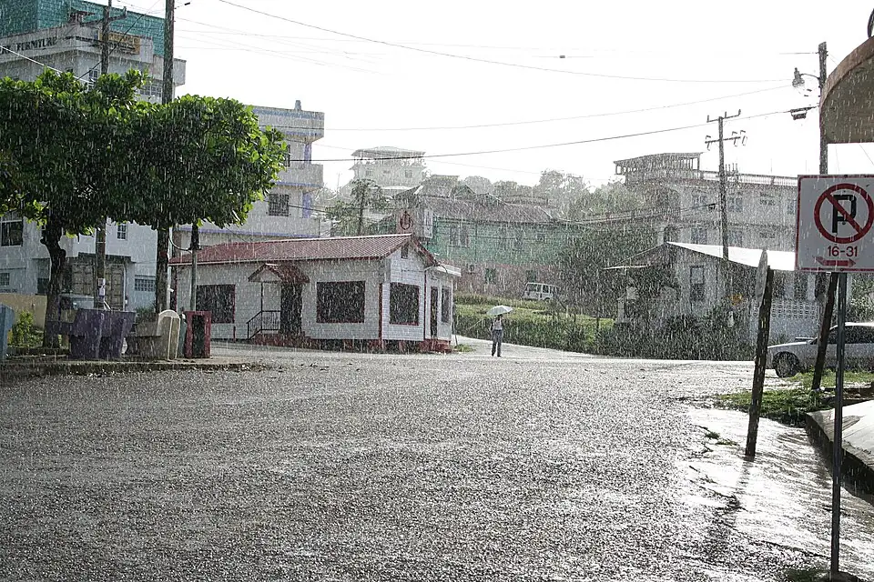 Heavy rain falling on an empty street corner in a hillside town, water running across the road and along the kerb, with a person under an umbrella in the distance.