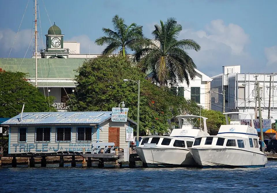 Two water-taxi boats moored beside a wooden marine terminal on the Belize City waterfront, with a clock tower and palms behind.