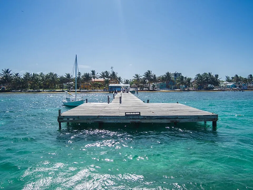 A wooden pier reaching out over turquoise water from the Caye Caulker shoreline, with a moored sailboat, coconut palms and painted village buildings along the shore behind it.