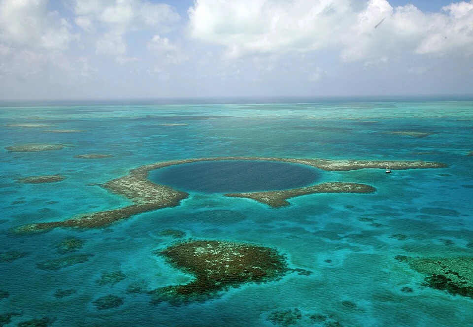 Aerial view of the Great Blue Hole, a dark circular sinkhole ringed by shallow turquoise reef, with a small boat on the water beside it.