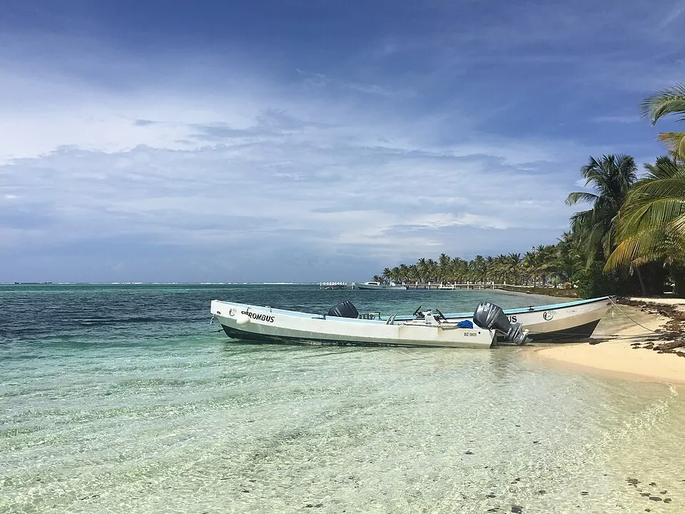 Two open dive boats pulled up on the white sand at Half Moon Caye, with a line of palms along the shore behind them.