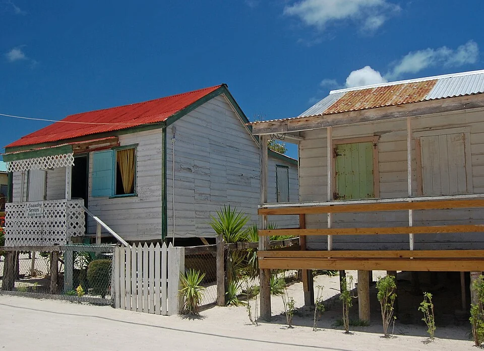 Two timber buildings standing on posts above the sand on Caye Caulker, one with a red tin roof and shuttered windows.