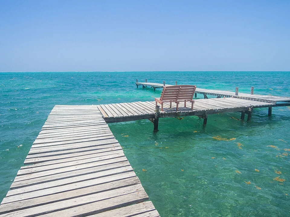 A weathered wooden pier with a single bench running out over shallow turquoise water off Caye Caulker under a clear sky.