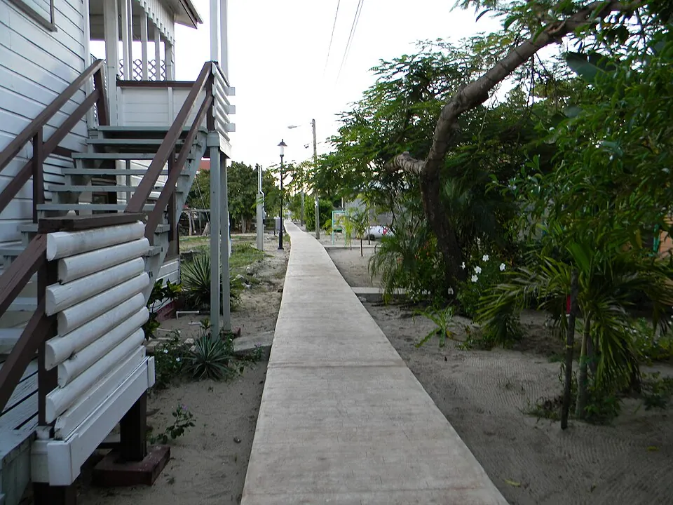 A narrow concrete walkway running between a timber building on stilts and sandy gardens in Placencia Village.